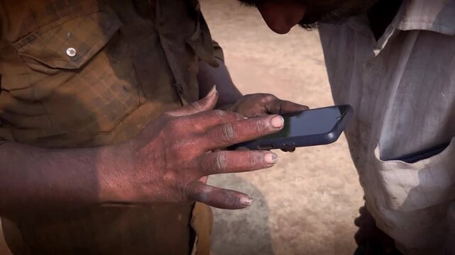 Two laborers with grimy hands peer at a smartphone with curiosity. The scene captures modernization as technology reaches the poor, bridging gaps and sparking a new digital era. No brand/no face.