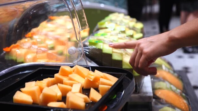 Woman's hand picking a piece of diced cantaloupe with a wooden stick from a fresh fruit display in a supermarket. The customer is choosing a healthy snack from a self service counter