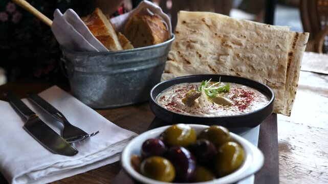 Close up of a woman's hand dipping a breadstick into a bowl of creamy baba ghanoush, served with pita bread, a bucket of rustic bread, and a side of mixed green and black olives on a wooden table