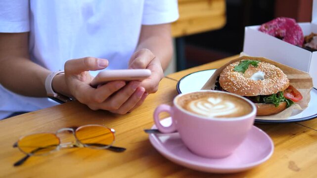 Anonymous woman sitting at a cafe table using her smartphone, with a pink cup of cappuccino, a fresh bagel sandwich, and sunglasses next to her, enjoying a modern breakfast outdoors