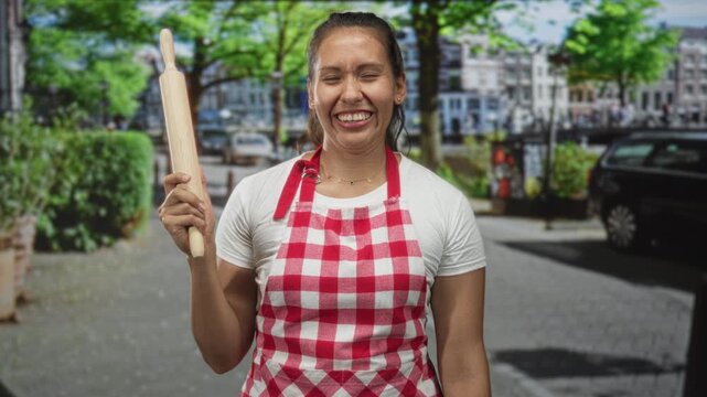 Woman cook smiling with eyes closed holds rolling pin upright on city street wearing red checkered apron and white tee; culinary joy.