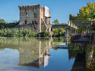 Obraz premium The Ruined Tower Of Scaliger Castle Reflecting In The Calm Mincio River Water Next To A Modern Bridge And Restaurant Area, Valeggio Sul Mincio, Italy