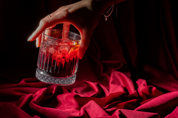 Hand holding a classic Negroni cocktail in a fluted glass, set against a luxurious, deep red draped fabric background with dramatic, moody lighting © pavel siamionov