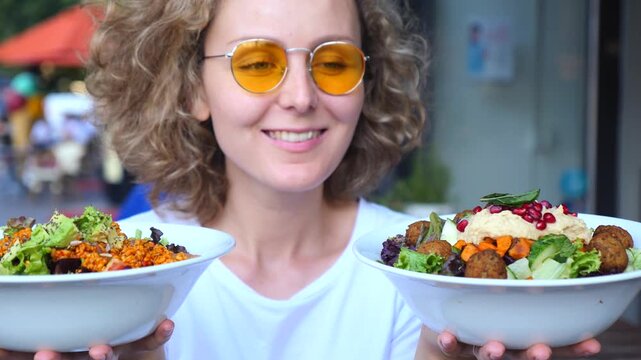 Young smiling woman with curly hair and sunglasses holding two bowls of healthy vegan food, including a falafel salad with hummus and a quinoa bowl, posing outdoors at a restaurant