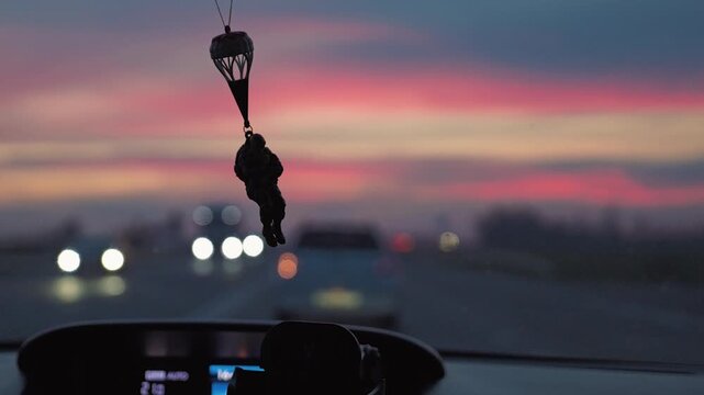 View from inside a car of a military paratrooper figurine. A colorful pink and purple sunset sky and blurry highway lights create a pensive, atmospheric mood. Travel and journey concept