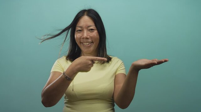 Woman pointing at open palm while presenting with both hands in studio with teal wall and wearing yellow shirt; friendly offering.