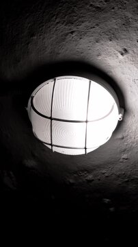 Old industrial bulkhead light with a protective cage flickers on a rough, dark concrete ceiling. Bright, cold light creates a stark, high-contrast and mysterious atmosphere.