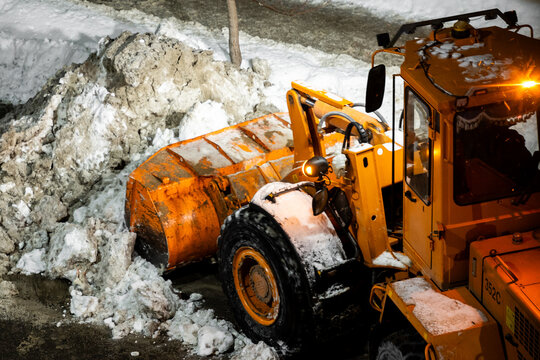An orange front-end loader clears a large pile of dirty snow at night under bright headlights.