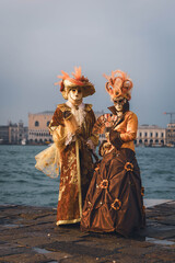 Fototapeta premium Venice, Italy - February 07, 2026: Two elegantly dressed individuals in ornate masks pose at Venice Carnival near the waterfront with historical buildings in the background