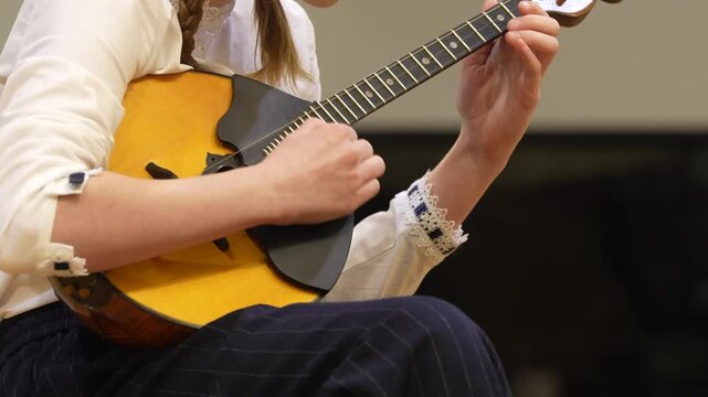 Female musician in white blouse plays a mandolin with intricate details, seated on a stage, showcasing her skill and focus during a musical performance