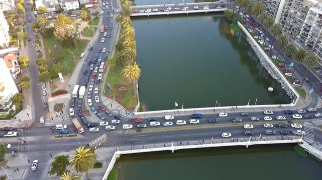 Aerial drone footage of urban traffic crossing a bridge over a city river with palm trees and surrounding buildings. Filmed above the Estero Marga Marga in Vina del Mar on a clear day.