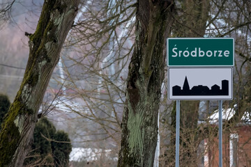 Tablica z nazwą wsi Śródborze, jak sama nazwa wskazuje, znajduje się w środku lasu, w zimowe popołudnie. Zielony znak drogowy wskazuje nazwę wsi, a na nim widnieje sylwetka miasta ( teren zabudowany) © Grzegorz