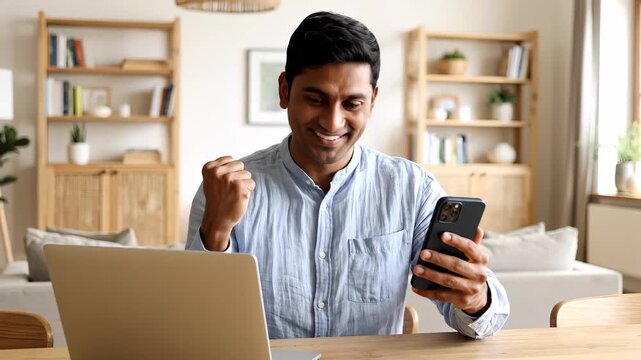 Excited indian man celebrating success or good news on smartphone while working from home with laptop