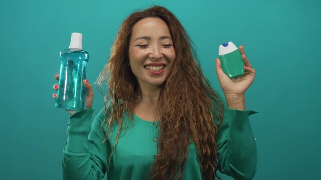 Woman smiling while holding a clear mouthwash bottle and green deodorant case in a teal studio, wearing a green sweater with long wavy hair visible; hygiene joy.