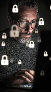 Male individual with gray beard and glasses focused on laptop screen displaying digital padlock icons, illustrating cybersecurity awareness in a dark environment