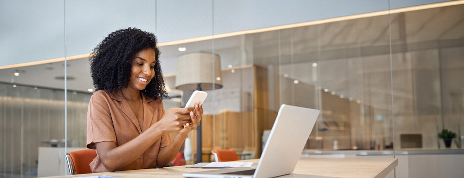 Happy young African American business woman holding smartphone sitting in office, smiling ethnic businesswoman using financial banking app on cell phone looking at cellphone working with mobile tech.