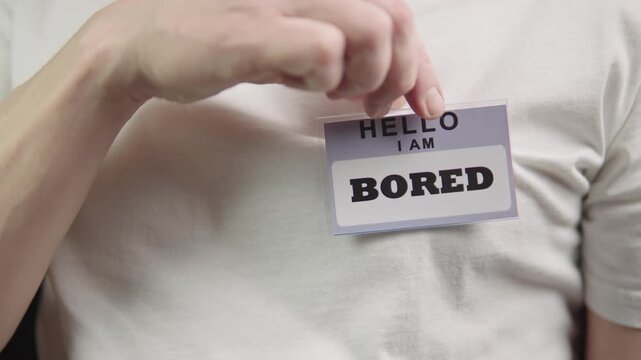 Individual holding bored badge name tag with dull expression casual attendee peels and places name tag reading bored