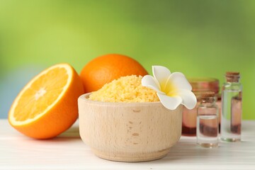 Yellow sea salt in bowl, oranges and flower on white wooden table against blurred green background,...