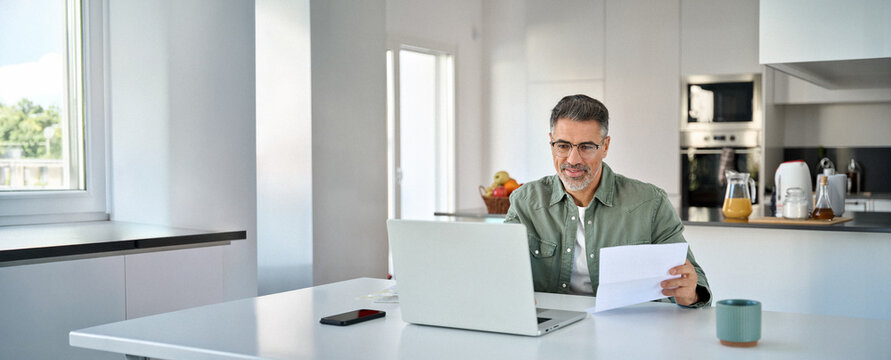Middle aged mature man senior customer wearing glasses looking at laptop computer holding paper bill checking financial invoice or tax document making online banking payments working at home table.
