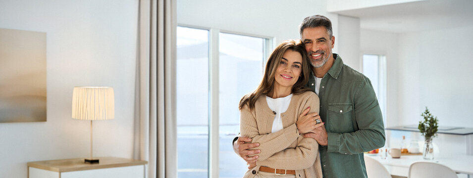 Smiling affectionate loving middle aged couple, happy romantic mature older Latin man and woman in their 50s hugging looking at camera standing together in modern kitchen at home. Portrait.