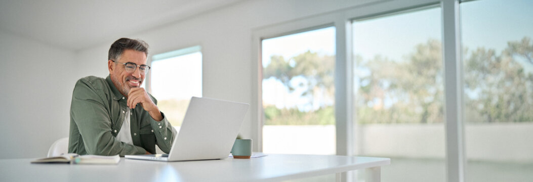 Smiling wearing eyeglasses looking at computer sitting at table. Happy older mature middle aged man using laptop hybrid working online, elearning, browsing web, searching online at home.