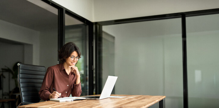 Smiling busy young professional businesswoman hr manager executive looking at laptop computer in office working sitting at desk. Woman employee having online meeting e-learning at work, writing notes.