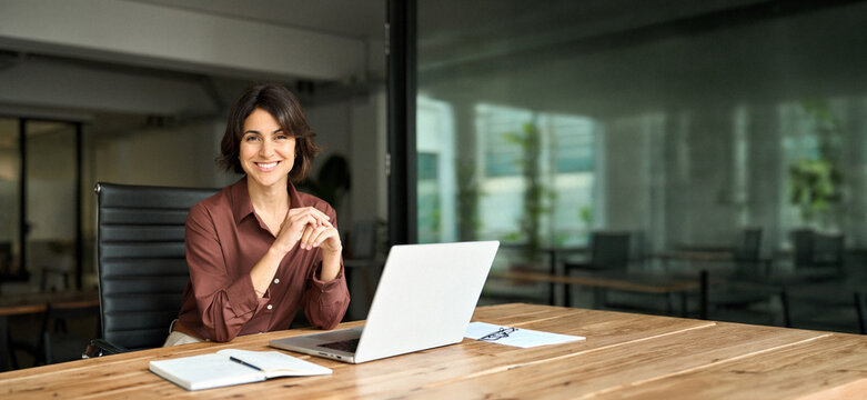 Smiling Hispanic young woman entrepreneur, happy female executive manager looking at camera sitting at workplace with laptop. Portrait of confident businesswoman leader in her 30s at work desk.