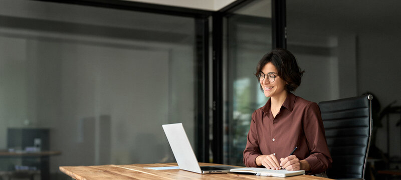 Female professional executive manager in her 30s using computer, elearning at workplace. Busy happy young Hispanic business woman company owner ceo working in office using laptop writing notes