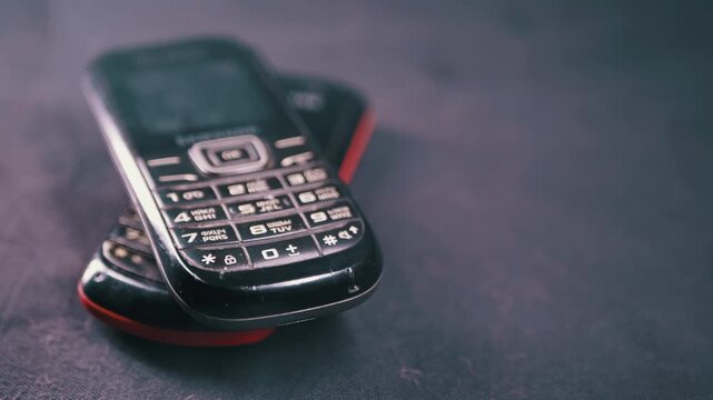 Close-up of two black vintage mobile phones with physical keypads stacked on a dark surface. Concept of obsolete technology, electronic waste, and the evolution of communication devices.