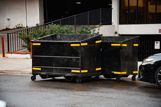Two large black industrial dumpsters with yellow safety stripes sit on a wet asphalt surface