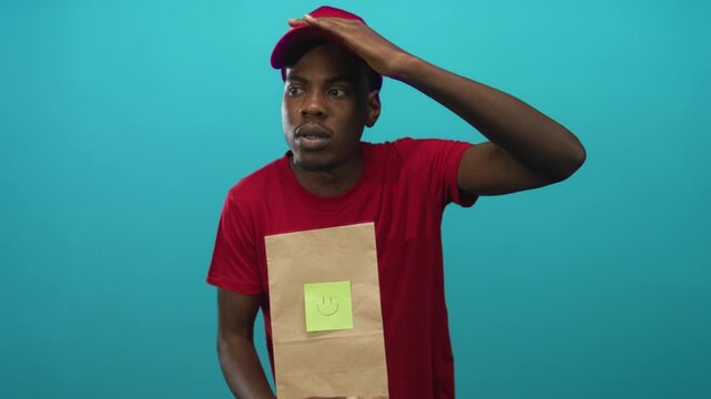 Young delivery man holds paper bag with smiley note and shades gaze with hand in studio; duty reliability.