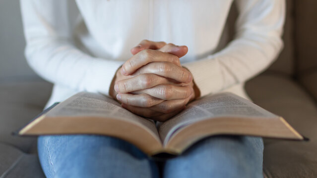 close up of woman praying with hands clasped over open bible at home, christian faith and devotion
