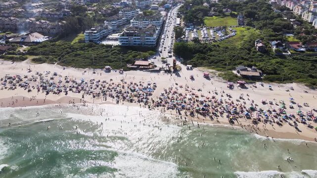High angle aerial view of a crowded Santinho Beach in Florianopolis, Brazil, showcasing seaside apartments, parking lots and people enjoying a sunny summer day by the ocean.
