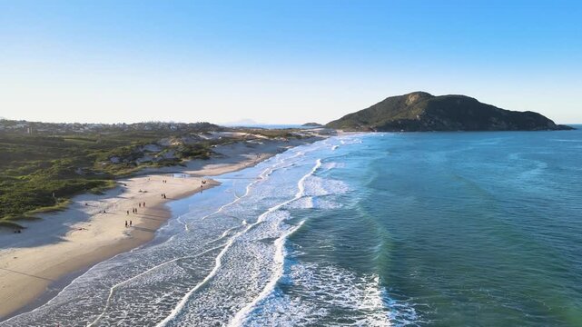 Cinematic aerial wide shot of Santinho Beach in Florianopolis, Brazil, featuring turquoise ocean waves, white sand dunes and the Morro dos Ingleses mountain under a clear blue sky.
