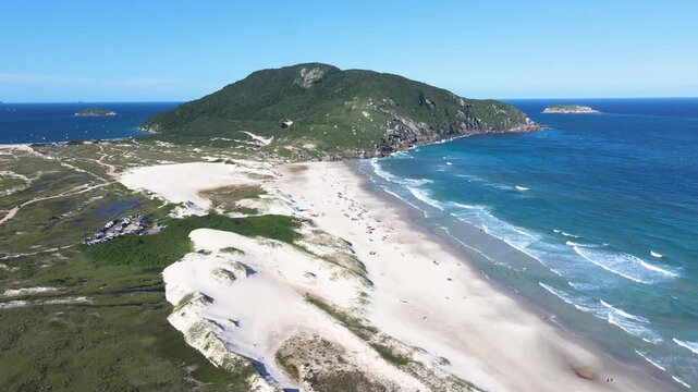 Stunning aerial panorama of Santinho Beach in Florianopolis, Brazil, showcasing the white sand dunes and the Morro dos Ingleses mountain under a clear blue summer sky.
