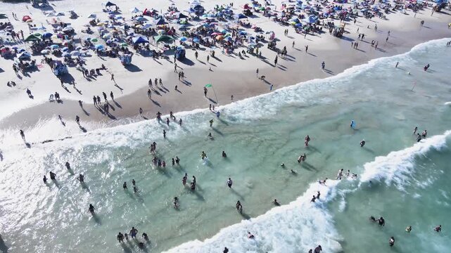 Birds eye view of a crowded Santinho Beach in Florianopolis, Brazil, with sun glinting on turquoise waves, people swimming and colorful umbrellas on the white sand.
