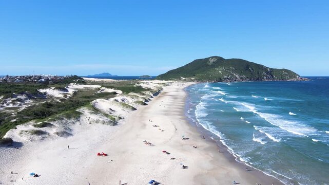Aerial wide shot of Santinho Beach in Florianopolis, Brazil, showcasing the white sand dunes and the Morro dos Ingleses mountain under a clear blue summer sky
