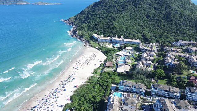Aerial landscape of Santinho Beach in Florianopolis, Brazil, featuring luxury resort buildings, crowded summer shoreline and green mountains under a bright sunny sky
