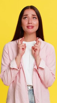 Young woman crossing fingers and closing eyes with hopeful expression, silently wishing for luck and good outcome. Girl on yellow background shows emotional anticipation and sincere desire for success