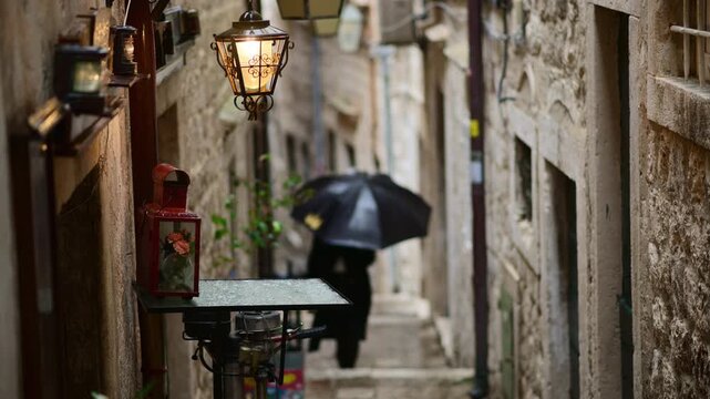 Person walking under an umbrella in a cobblestone alley with stone walls and vintage lanterns