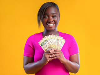 A smiling woman in a bright pink shirt stands against a vibrant yellow background. She is proudly fanning out a stack of 5,000 Jamaican dollar banknotes, looking directly at the camera with joy. © RODWORKS