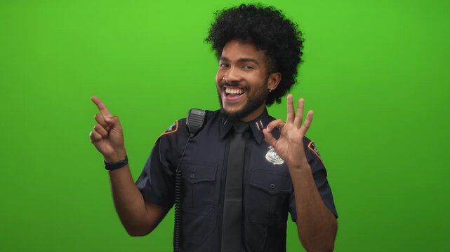 Smiling man in police uniform with green background showing okay sign and pointing, creating a positive, cheerful atmosphere.