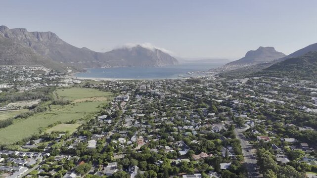 Drone facing south, flies to the right over city with vew of Chapman's Peak covered in clouds on a sunny morning in Hout Bay, Cape Town, South Africa