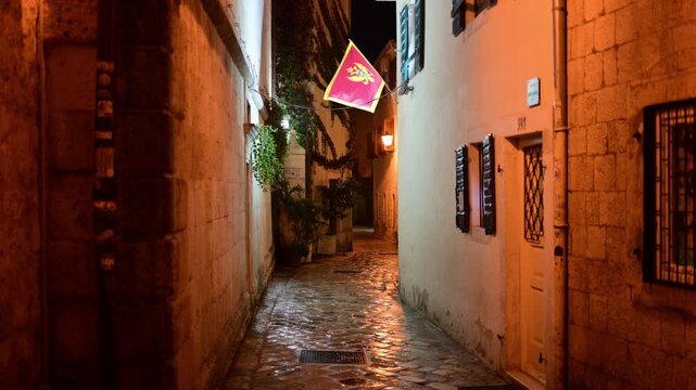 Montenegro flag hanging in a traditional narrow stone alley with cobblestone street and warm night lighting. Kotor Old Town