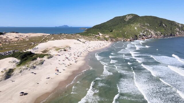Cinematic aerial wide shot of Santinho Beach in Florianopolis, Brazil, featuring white sand dunes, turquoise ocean waves and the Morro dos Ingleses mountain under a clear blue sky
