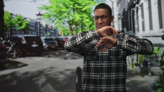 Young black man shielding his face with crossed hands on a city street, wearing glasses and a flannel shirt; discomfort avoidance privacy.