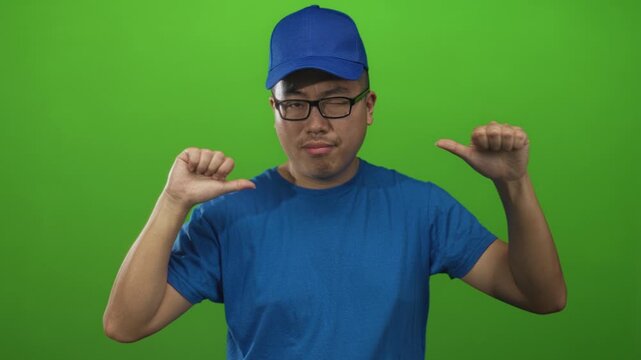 Man with blue cap and glasses pointing thumbs to chest in green studio setting while winking; confident attitude.