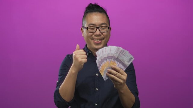 Young man holding a fanned stack of indonesian rupiah bills and giving thumbs up while smiling in a purple studio; celebration success.