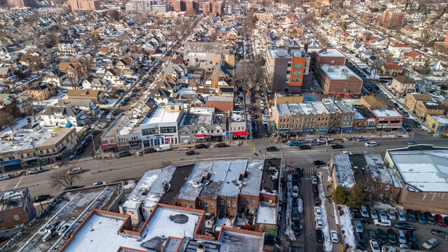 Aerial view capturing a new york city neighborhood street with a mix of residential homes and commercial buildings, featuring vehicles navigating roads and snow dusted rooftops during winter
