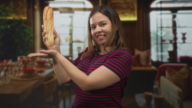 Young woman holding a baguette with palm up presenting gesture in hotel dining room near table and chairs; playful moment.